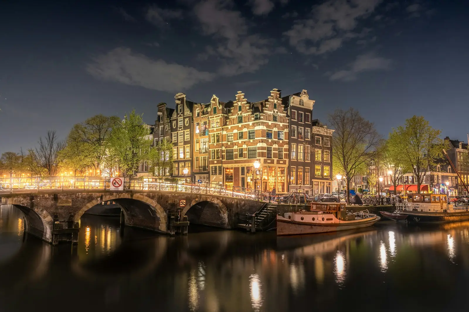 Verlichte grachtenpanden achter een stenen boogbrug over een Amsterdamse gracht bij avond