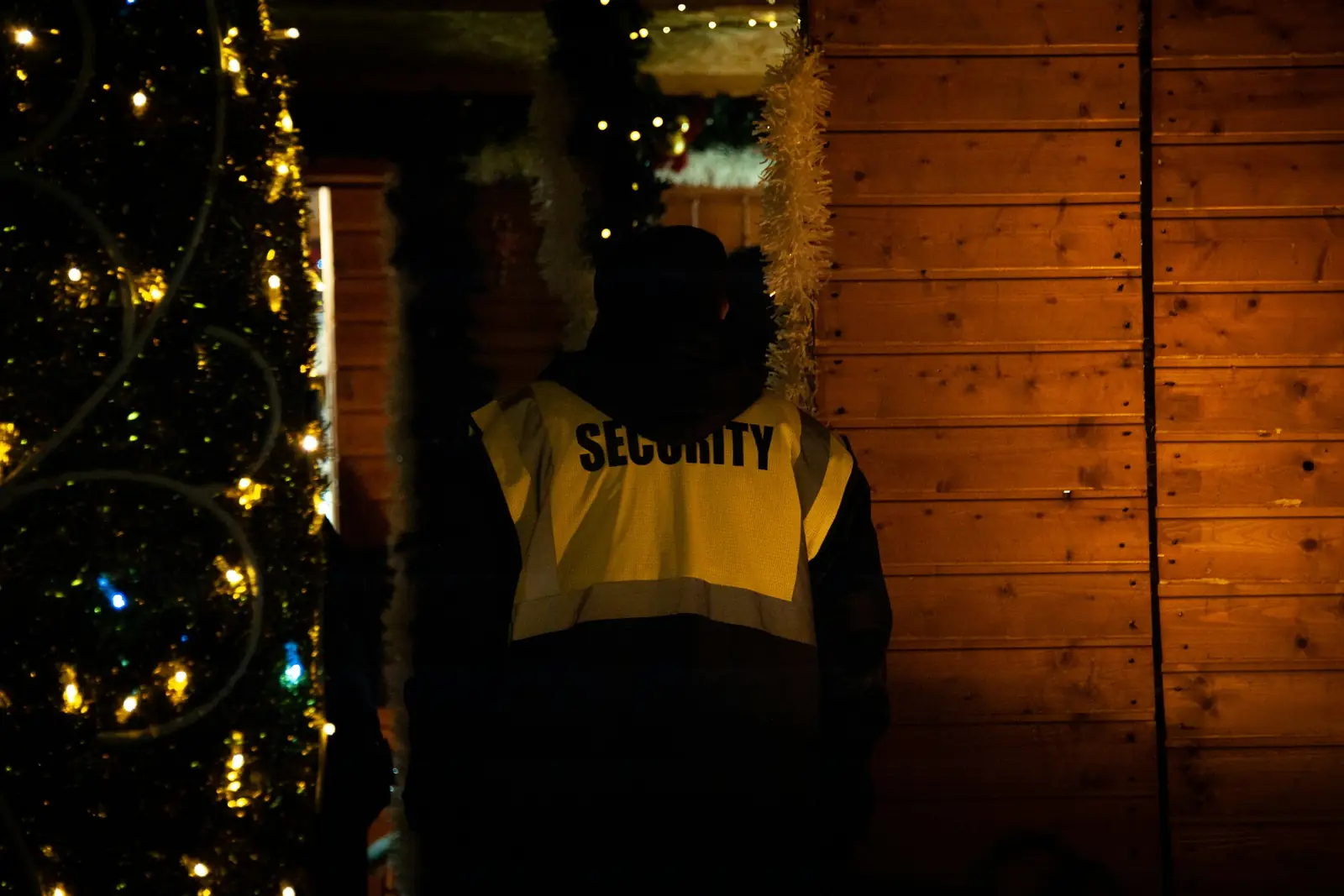 Beveiliger in geel hesje, van achteren gezien, naast een verlichte kerstboom op een avondmarkt